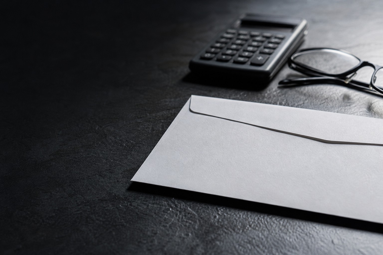 Close-up of a blank sealed envelope, a calculator, and reading glasses on a dark slate surface.