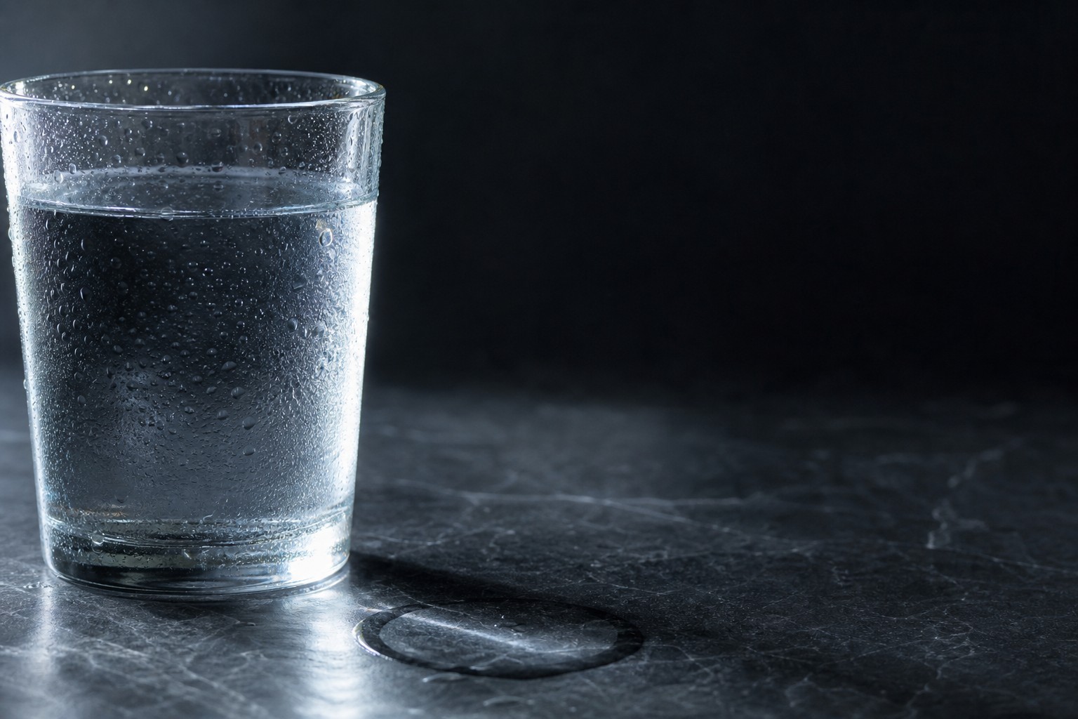 Close-up of a clear glass of water on a dark stone counter with condensation catching low side light.